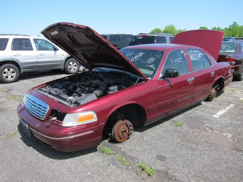2001 Ford Crown Victoria - A red 2001 Ford Crown Victoria sits in a junkyard with its hood open and all four wheels removed.