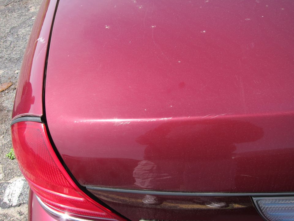 Close-up of a red car's trunk lid and taillight. Shows scratches and scuffs on the paint