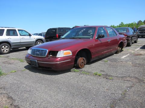 2001 Ford Crown Victoria - A maroon 2001 Ford Crown Victoria sits without wheels in a junkyard lot.