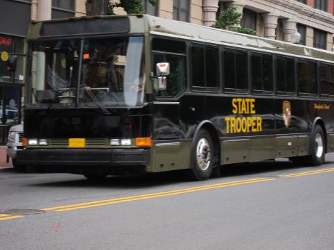 1991 Ford LTD Crown Victoria - black State Trooper bus with gold lettering moving along an urban street lined with buildings.