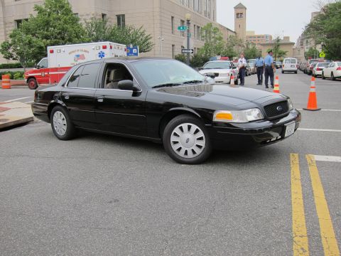 1991 Ford LTD Crown Victoria - unmarked black Ford Crown Victoria sedan on a city street, with an ambulance and uniformed officers in the background.