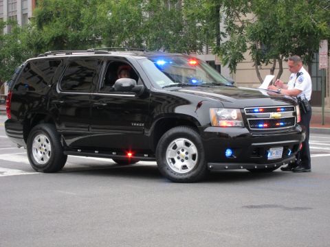 1991 Ford LTD Crown Victoria - black Chevrolet Tahoe police SUV with grille lights and an officer writing at the front fender on a city intersection.