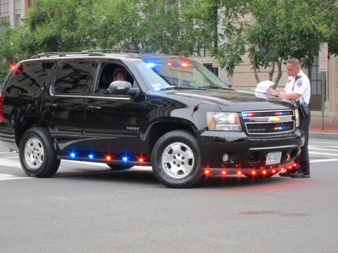 1991 Ford LTD Crown Victoria - black Chevrolet Tahoe police SUV with red and blue emergency lights illuminated and an officer standing at its front.