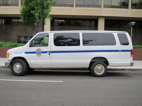 1991 Ford LTD Crown Victoria - Driver-side profile of a white Ford Econoline police passenger van with a blue stripe parked at a city curb.