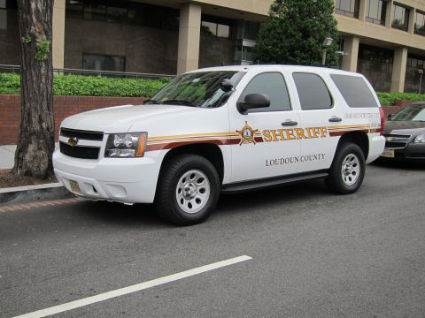 1991 Ford LTD Crown Victoria - white Chevrolet Tahoe sheriff SUV marked Loudoun County parked at a curb beside a brick wall and tree.