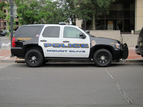 1991 Ford LTD Crown Victoria - Side profile of a black-and-white Mount Olive Police Chevrolet Tahoe with push bar parked along a curb.