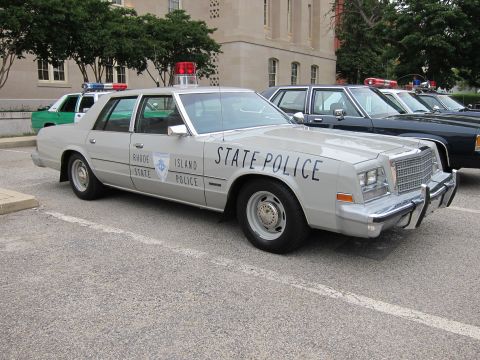 1991 Ford LTD Crown Victoria - silver vintage Chrysler-style Rhode Island State Police sedan parked among other police cars by a stone building.