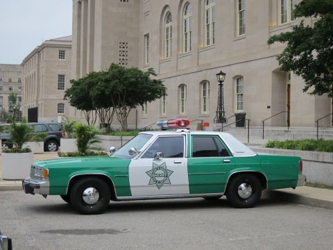 1991 Ford LTD Crown Victoria - Side profile of a green-and-white Ford Crown Victoria sheriff car with star emblem parked near a stone building.