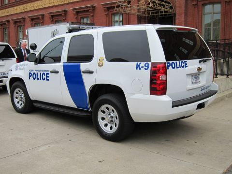 1991 Ford LTD Crown Victoria - white Chevrolet Tahoe marked Federal Protective Service Police K-9 with blue stripe, parked by a brick building.