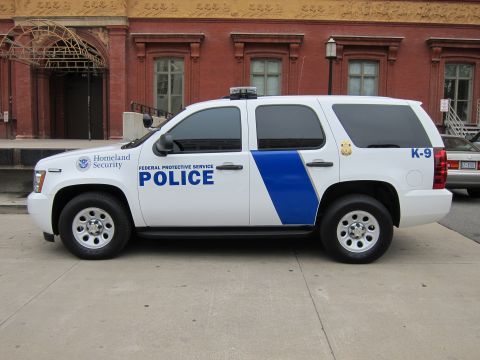 1991 Ford LTD Crown Victoria - white Chevrolet Tahoe police SUV marked Homeland Security Federal Protective Service Police, parked along a brick building.
