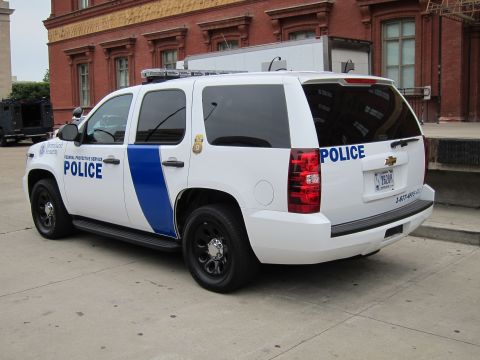 1991 Ford LTD Crown Victoria - white Chevrolet Tahoe marked Federal Protective Service Police with blue stripe, parked near a brick building and an armored vehicle.