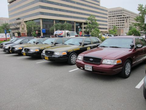 1991 Ford LTD Crown Victoria - lineup of olive-green State Trooper Ford sedans angled in a city lot, with a maroon Ford Crown Victoria parked alongside.