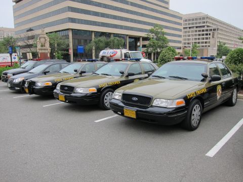1991 Ford LTD Crown Victoria - Several olive-green Ford State Trooper sedans lined up at an angle in a downtown plaza lot with office buildings behind.