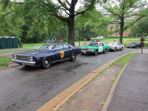 1991 Ford LTD Crown Victoria - row of vintage police cars on a wet park road, led by a dark-blue Plymouth sedan followed by green-and-white cruisers.