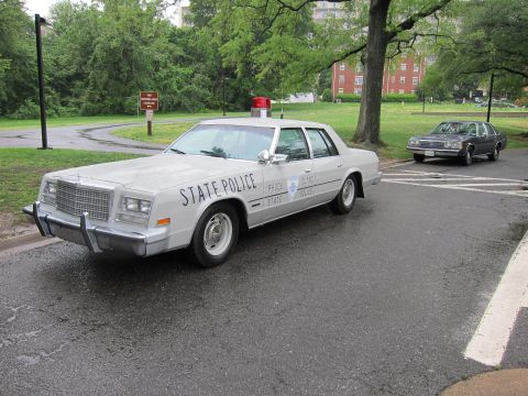 1991 Ford LTD Crown Victoria - silver vintage State Police sedan with a single red beacon parked on a park road, with another classic car behind.