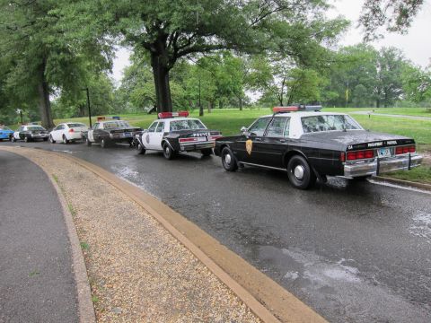 1991 Ford LTD Crown Victoria - lineup of classic black-and-white police and highway patrol sedans parked along a wet curving road in a green park.