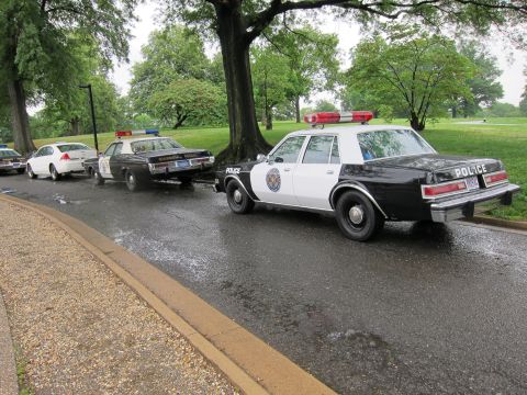 1991 Ford LTD Crown Victoria - black-and-white Police cruiser with a roof light bar parked behind other classic patrol cars on a rain-slicked park road.
