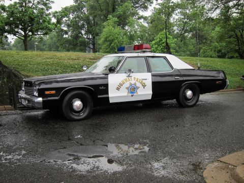 1991 Ford LTD Crown Victoria - classic black-and-white Highway Patrol sedan with front push bar parked beside a grassy embankment on a wet road.