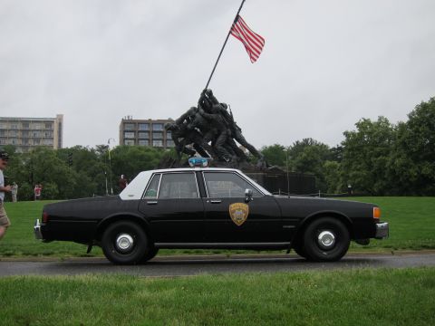 1991 Ford LTD Crown Victoria - black police sedan parked on grass in front of the Iwo Jima Marine Corps War Memorial flying a US flag.