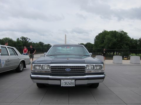 1991 Ford LTD Crown Victoria - Head-on view of a black Ford four-door sedan on a plaza, the Washington Monument centered behind it under gray skies.