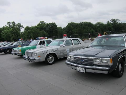 1991 Ford LTD Crown Victoria - row of vintage police cars including a green State Police sedan and a black Ford, lined up on a stone plaza.