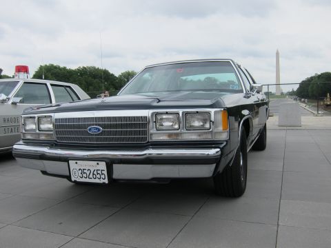 1991 Ford LTD Crown Victoria - Front three-quarter view of a black Ford four-door sedan on a plaza with the Washington Monument and trees behind.