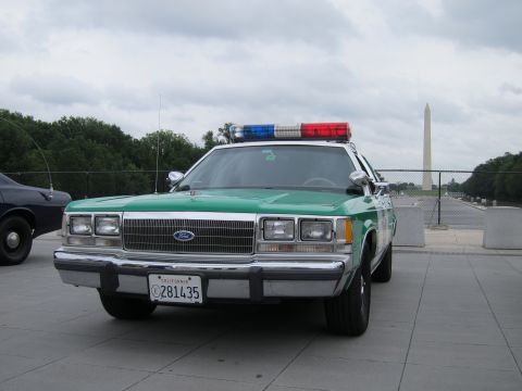 1991 Ford LTD Crown Victoria - Front three-quarter view of a green-and-white Ford police sedan with roof lightbar and California plate, Washington Monument behind.