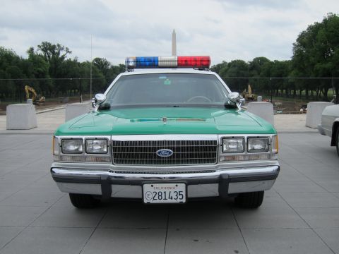 1991 Ford LTD Crown Victoria - Head-on view of a green-and-white Ford police sedan with red-and-blue roof lightbar, California plate, Washington Monument behind.