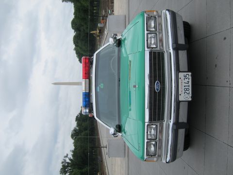 1991 Ford LTD Crown Victoria - Vertical head-on view of a green-and-white Ford police sedan with a lightbar, the Washington Monument centered behind it.