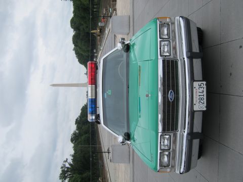 1991 Ford LTD Crown Victoria - Head-on view of a green-and-white Ford police sedan with roof lightbar and California plate, Washington Monument in the background.