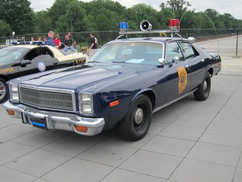 1991 Ford LTD Crown Victoria - dark blue police sedan with a gold door emblem, roof lightbar and spotlight parked on a plaza, spectators behind.
