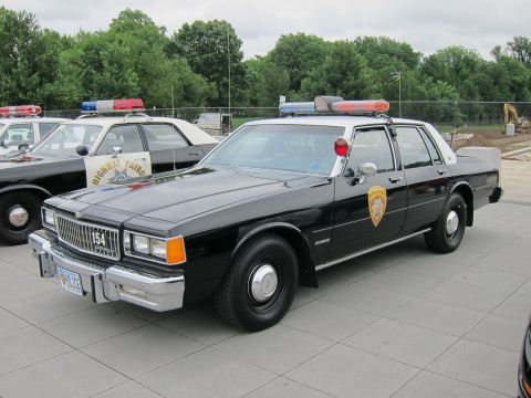 1991 Ford LTD Crown Victoria - black-and-white police sedan numbered 154 with a roof lightbar and door shield, parked on a stone plaza among other cars.