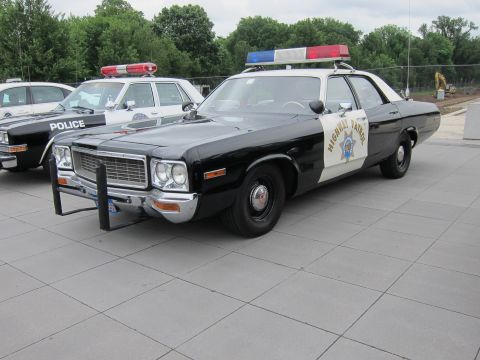 1991 Ford LTD Crown Victoria - Front three-quarter view of a black-and-white Highway Patrol sedan with roof beacons, parked on a plaza with trees behind.