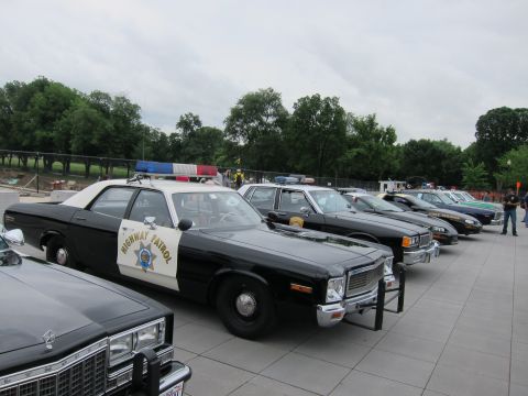 1991 Ford LTD Crown Victoria - long row of vintage police cars led by a black-and-white Highway Patrol sedan, parked diagonally on a stone plaza.