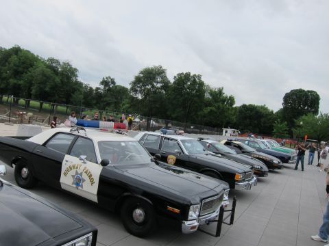 1991 Ford LTD Crown Victoria - line of classic police cars led by a Highway Patrol sedan, with spectators walking nearby on a tree-lined plaza.