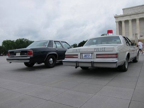 1991 Ford LTD Crown Victoria - dark vintage sedan beside a white STATE POLICE cruiser with red light bar on a paved plaza near a memorial.