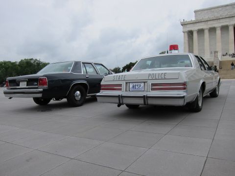 1991 Ford LTD Crown Victoria - Rear view of a black sedan and a white STATE POLICE cruiser parked together on a stone plaza under gray skies.