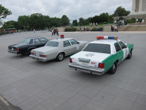 1991 Ford LTD Crown Victoria - Three vintage police cruisers, including a green-and-white SHERIFF Crown Victoria, parked at an angle on a plaza.