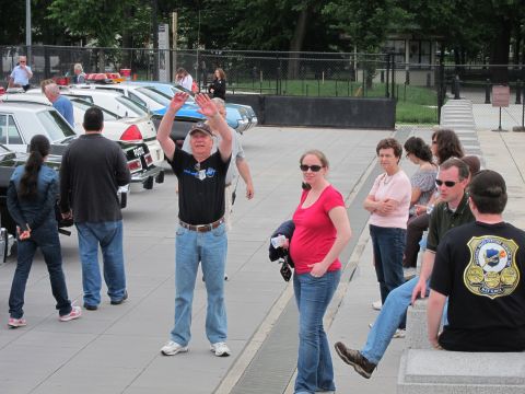 1991 Ford LTD Crown Victoria - man in a cap waves while standing among visitors at an outdoor gathering with police cars lined up behind.