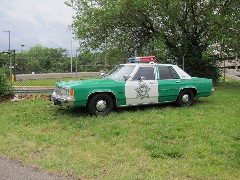 1991 Ford LTD Crown Victoria - green-and-white SHERIFF Ford LTD Crown Victoria with light bar parked on grass near a chain-link fence.