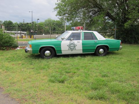1991 Ford LTD Crown Victoria - Side profile of a green-and-white SHERIFF Ford sedan with light bar parked on a grassy lot by a fence.
