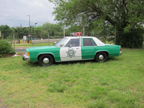 1991 Ford LTD Crown Victoria - two-tone green and white sheriff police sedan with roof light bar parked on grass near a fence.