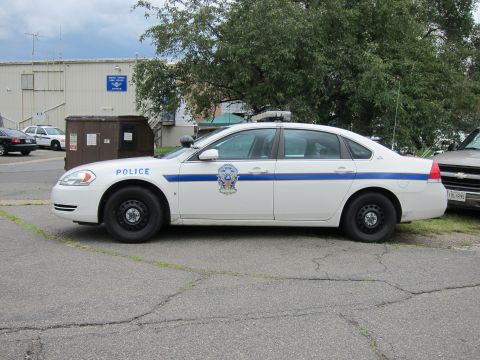 1991 Ford LTD Crown Victoria - white police sedan marked Police with a blue stripe parked on pavement beside a dumpster and building.