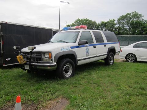 1991 Ford LTD Crown Victoria - white Chevrolet Suburban police SUV with blue stripe and roof light bar parked on grass by a trailer.