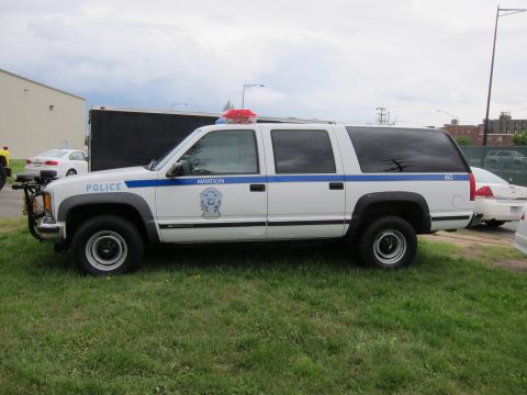 1991 Ford LTD Crown Victoria - white and gray Chevrolet Suburban police SUV with blue stripe and roof lights shown in side profile on grass.