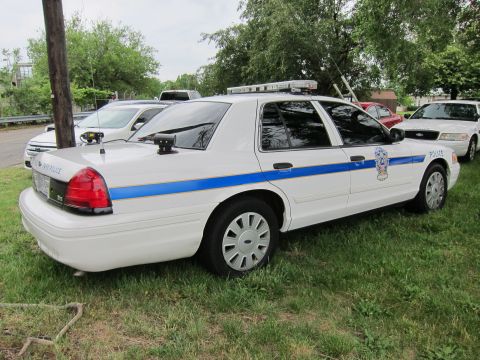 1991 Ford LTD Crown Victoria - white Ford Crown Victoria police sedan with blue stripe and roof light bar parked on grass.