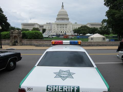 1991 Ford LTD Crown Victoria - Rear roof and red-blue lightbar of a green-and-white Sheriff car with the U.S. Capitol and event tents in the background.