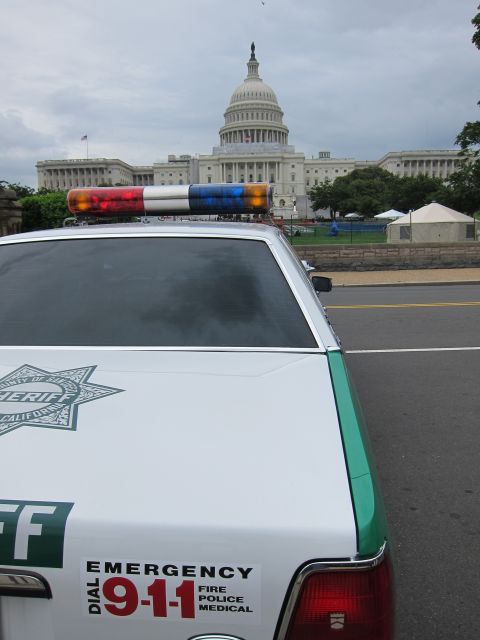1991 Ford LTD Crown Victoria - Side-rear view of a white-and-green Sheriff patrol car with an EMERGENCY 9-1-1 decal, the U.S. Capitol in the distance.
