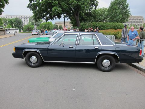 1991 Ford LTD Crown Victoria - dark blue four-door sedan shown in side profile at an outdoor gathering with other cars and spectators behind.