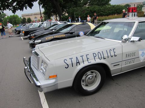 1991 Ford LTD Crown Victoria - Front quarter of a silver Rhode Island State Police sedan with a black State Trooper car and lineup behind it.